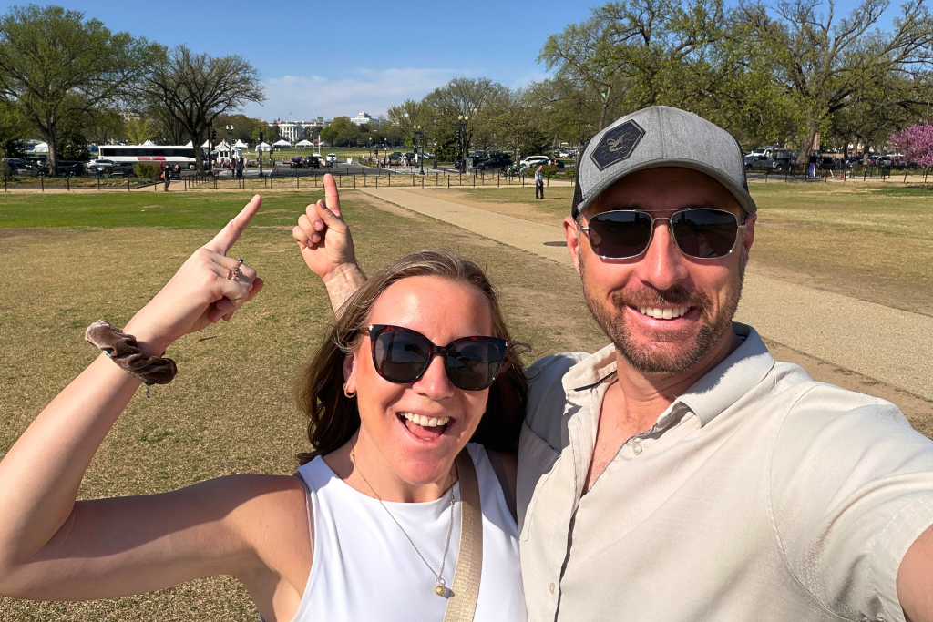 Kate and her husband smile and point toward the White House in the distance while standing on the National Mall during a sunny day exploring Washington DC.