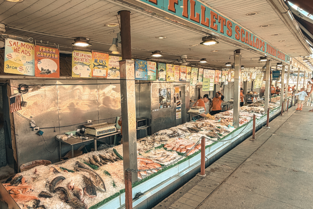Fresh seafood displayed on ice at an outdoor fish market in Washington DC with signs above reading items like "Fresh Catfish" and "Salmon Heads" as people shop along the counter.