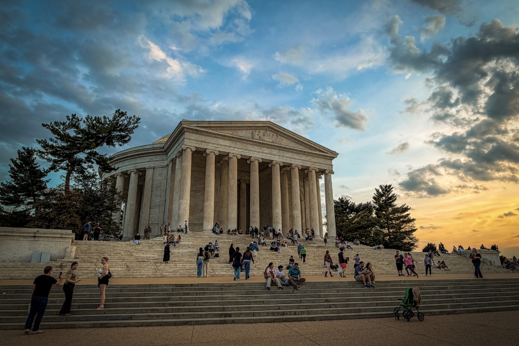 The Jefferson Memorial in Washington DC at sunset with people sitting on the steps and a golden sky with clouds overhead.