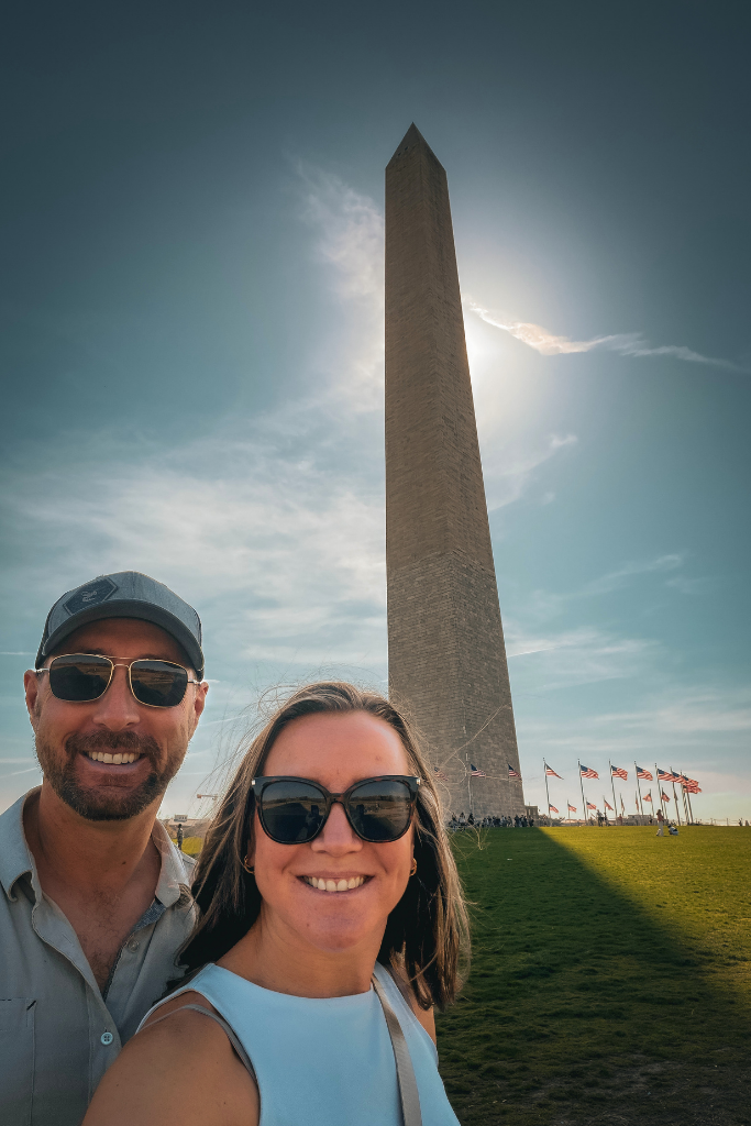 Kate and her husband smile while taking a selfy in front of the Washington Monument with flags waving on a sunny afternoon.