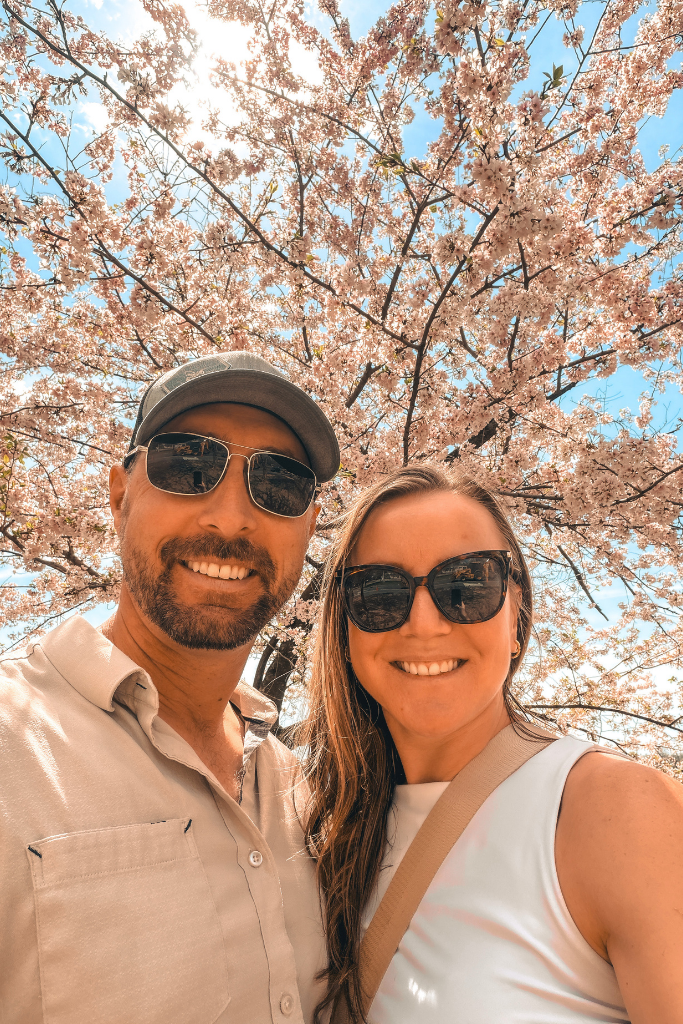 Kate and her husband smile together under blooming cherry blossom trees on a sunny spring day in Washington DC.