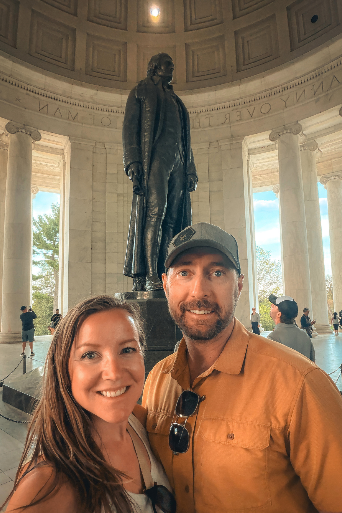 Kate and her husband smile for a selfie inside the Jefferson Memorial with the statue of Thomas Jefferson standing behind them.