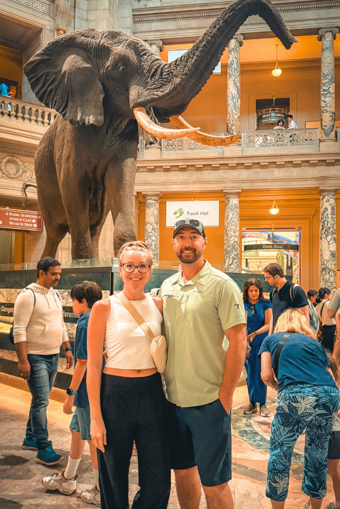 Kate and her husband are smiling in front of a large elephant display inside a natural history museum, with a sign reading "Fossil Hall" visible behind them.