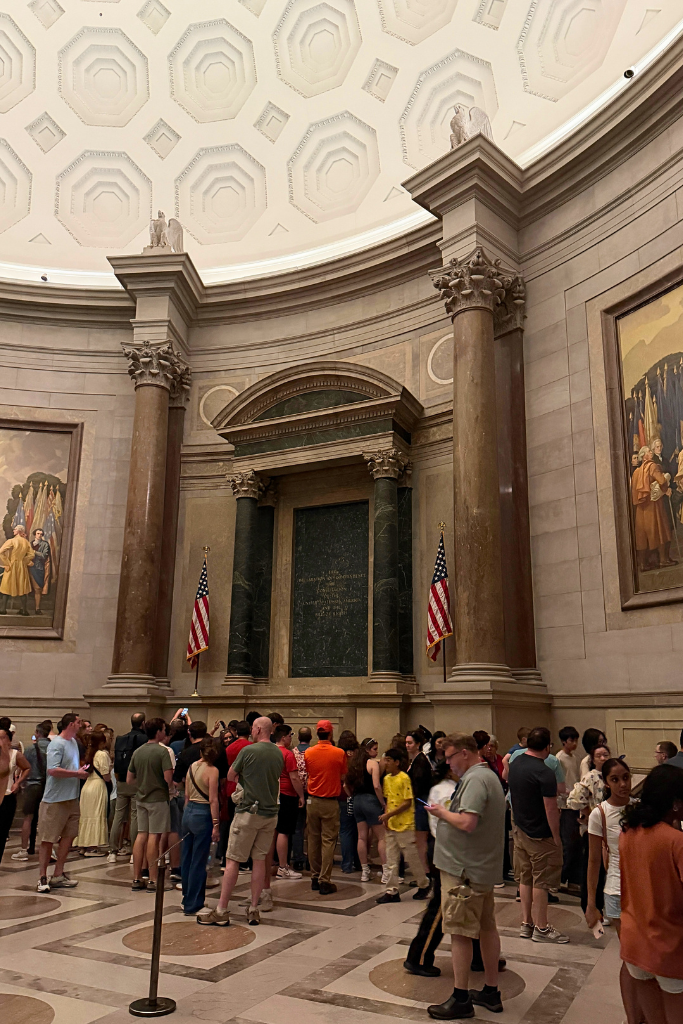 Visitors gathered inside a grand rotunda with a patterned domed ceiling and large columns, viewing historic documents displayed between two American flags in the Archives Museum.