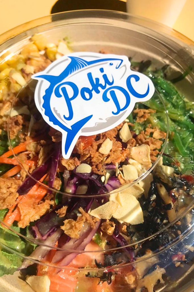 Fresh poke bowl with salmon, seaweed salad, shredded carrots, red cabbage, crispy garlic chips, and crunchy toppings in a clear container labeled "Poki DC" on the lid.