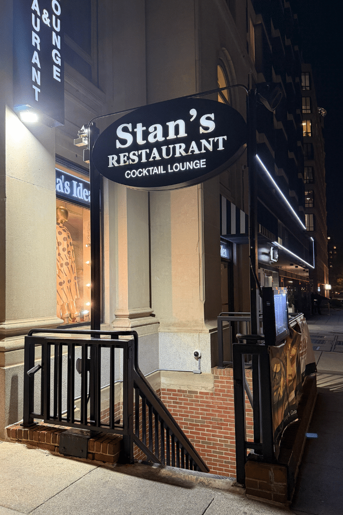 Nighttime street view in Washington DC with a glowing sign that reads "Stan’s Restaurant Cocktail Lounge" above a stairway entrance along a city sidewalk.