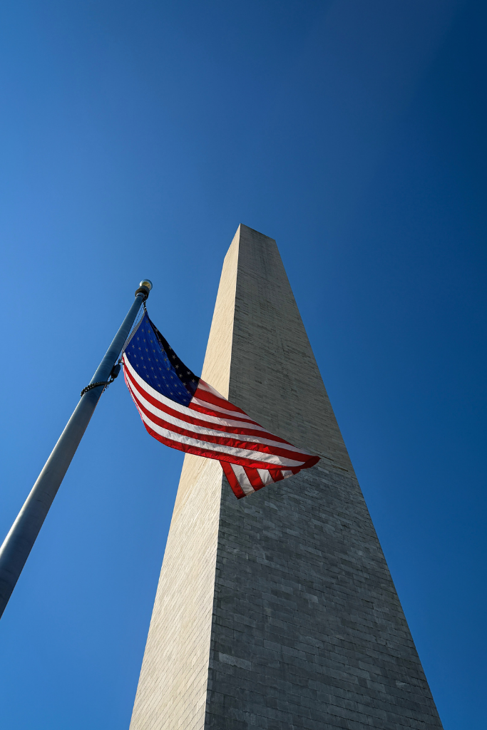 The Washington Monument rises into a clear blue sky with an American flag waving in the foreground.