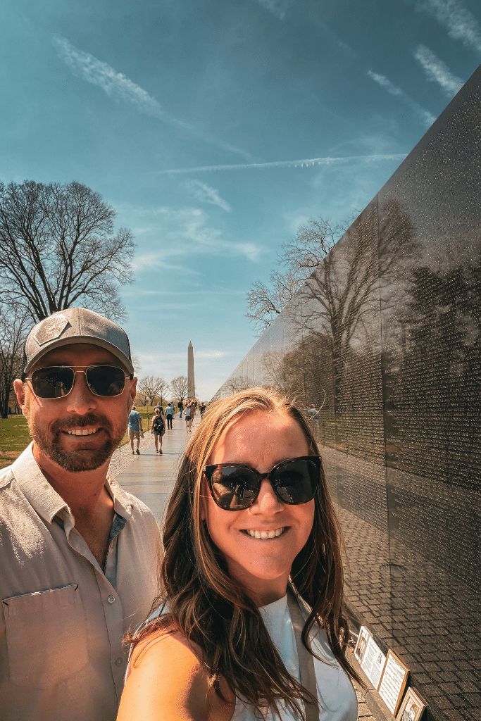 Kate and her husband smile for a selfie beside the Vietnam Veterans Memorial wall in Washington DC with the Washington Monument visible in the distance on a sunny day exploring the city
