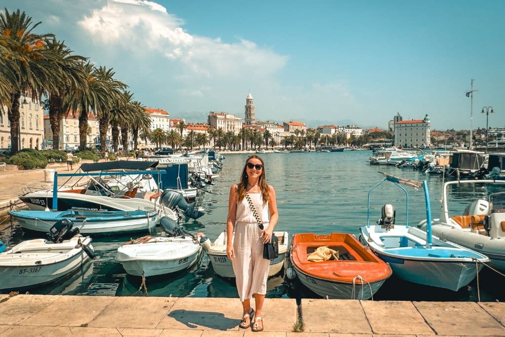 Kate stands on the edge of a dock in the marina filled with boats and the shoreline of Old Town Split in the background on a sunny day.