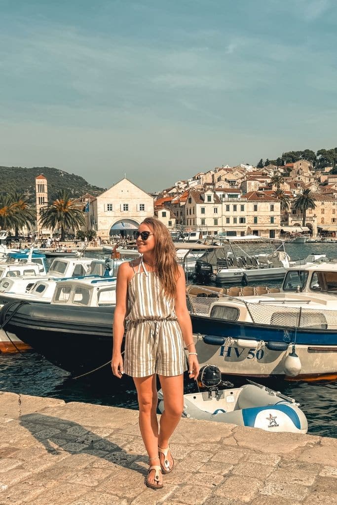 Kate is standing, smiling along the shore in Hvar with the marina and stunning coastline in the background.