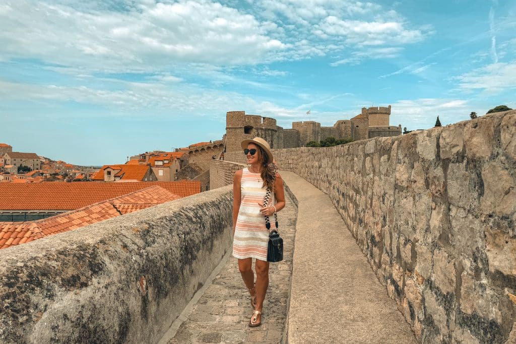Kate walking alone along the top of the wall surrounding Dubrovnik, wearing a pink and white stripe sundress.