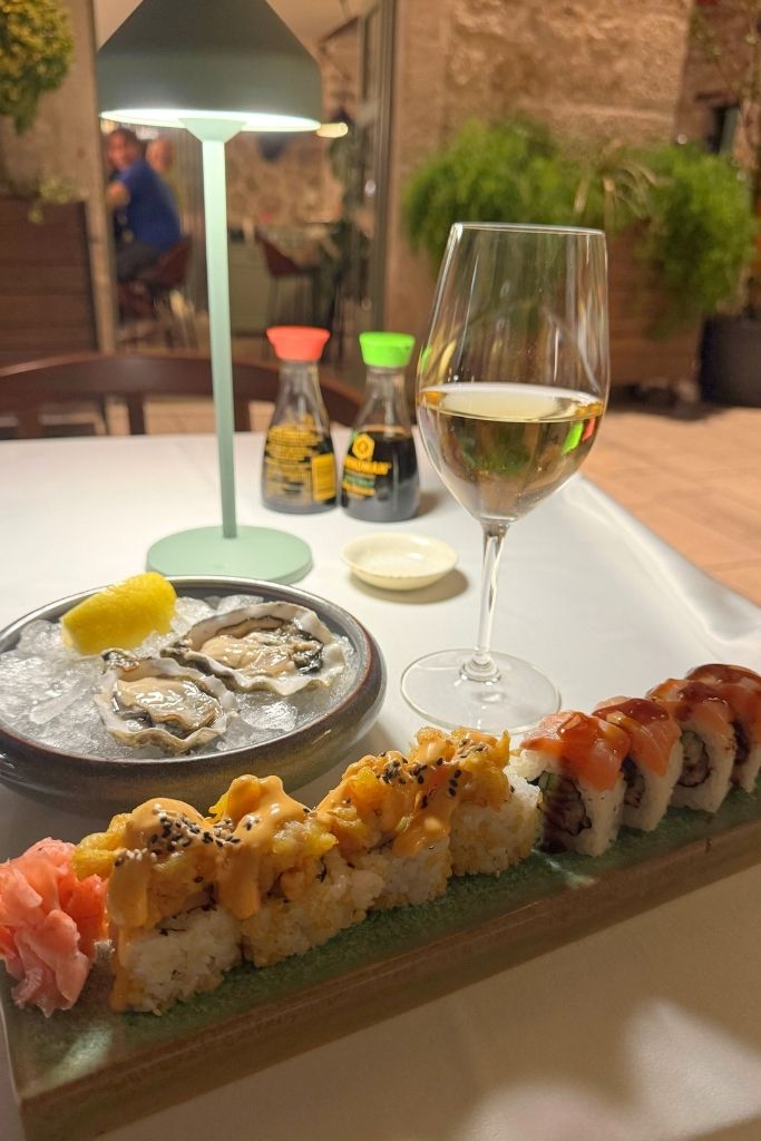 A glass of wine with a plate of raw oysters and a sushi roll on a white linen tablecloth.