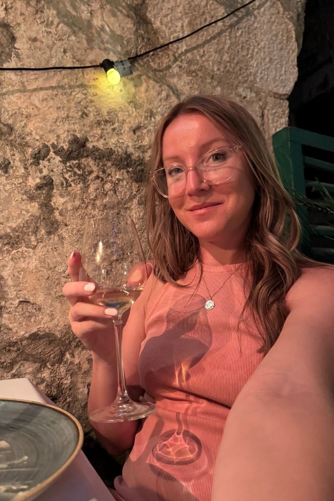 Kate is taking a selfie holding a glass of white wine under candlelight. 