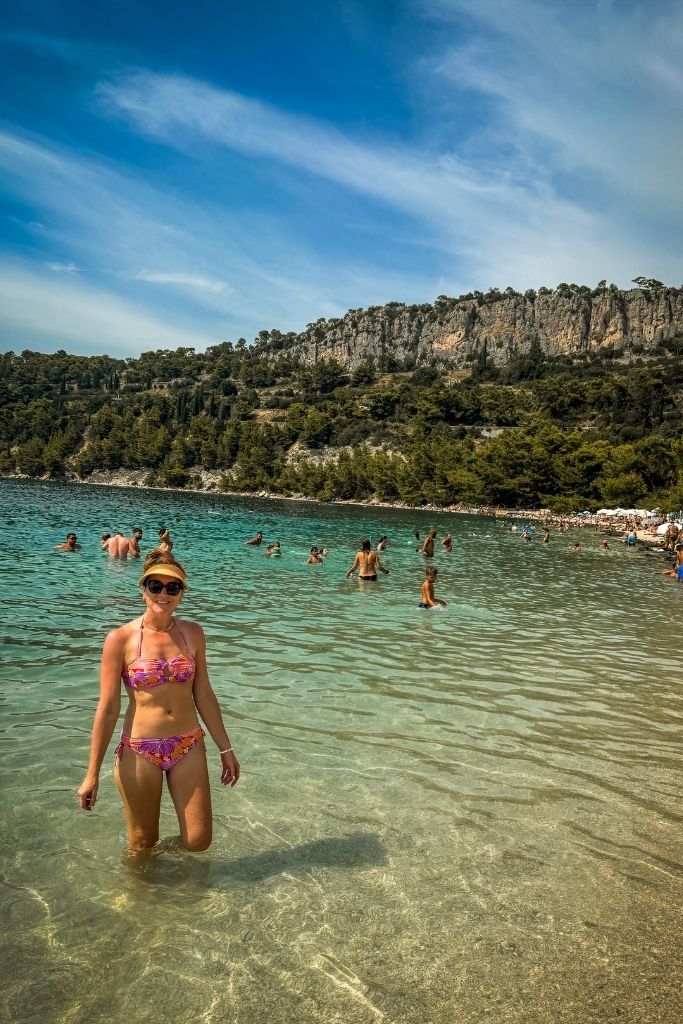 Kate is wading in the crystal clear blue sea along the Croatian coast, on a sunny day, wearing a pink swimsuit.