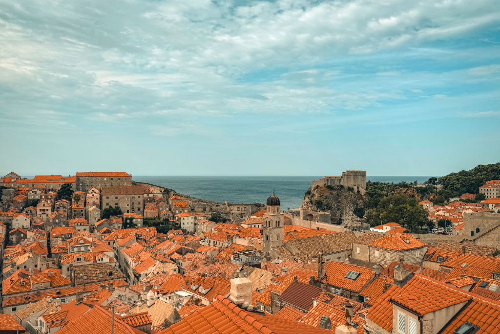 A view of Dubrovnik's orange rooftop buildings surrounded by the stone wall and the sea in the distance.  
