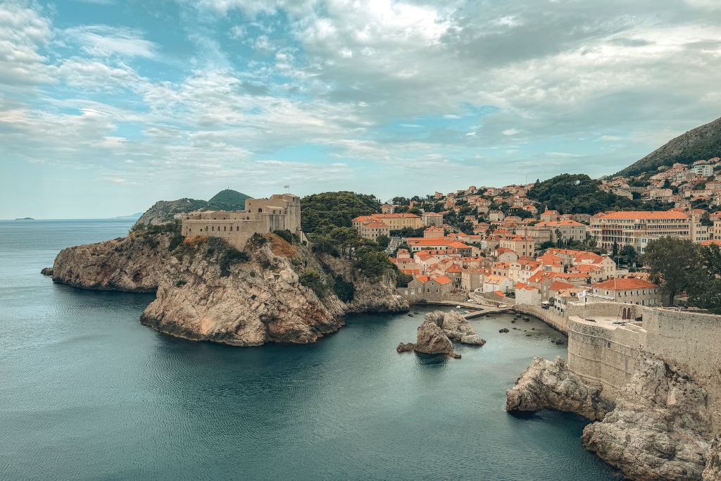 View of Croatia's rocky coastline with an old fort on top of a cliff surrounded by orange-roofed buildings below.