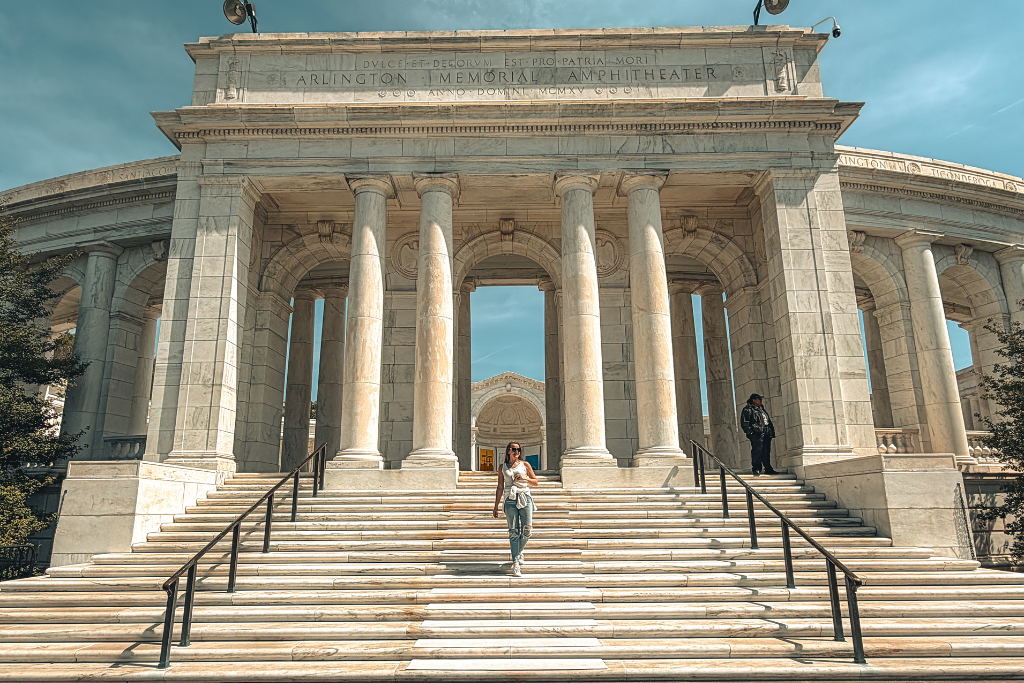 Kate walking down the steps of Arlington Memorial Amphitheater framed by tall marble columns with engraved text "Arlington Memorial Amphitheater" visible above the entrance at Arlington National Cemetery.