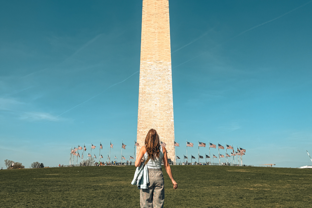 Kate walking toward the Washington Monument off in the distance that towers into a blue sky, surrounded by American flags at the base.