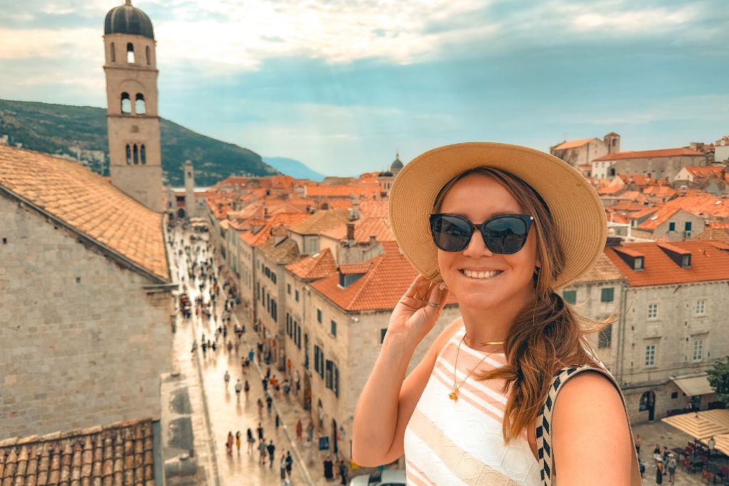Kate taking a selfie from top the wall in Dubrovnik with old town in the background. She's smiling and holding onto her sunhat.