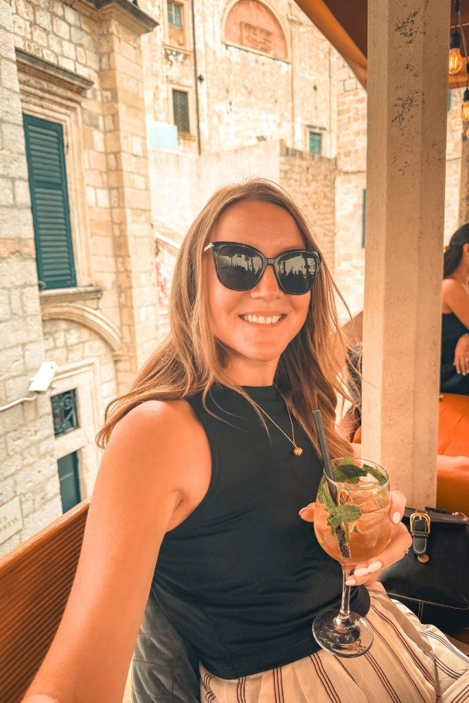 Kate taking a selfie with a Hugo spritz in her hand, smiling at the camera. Old stone walls behind her. 