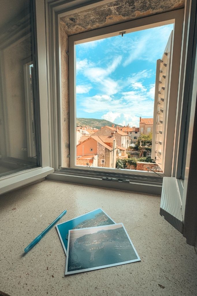 Open window with a view of Dubrovnik's red roof buildings and two postcards on the window seal.