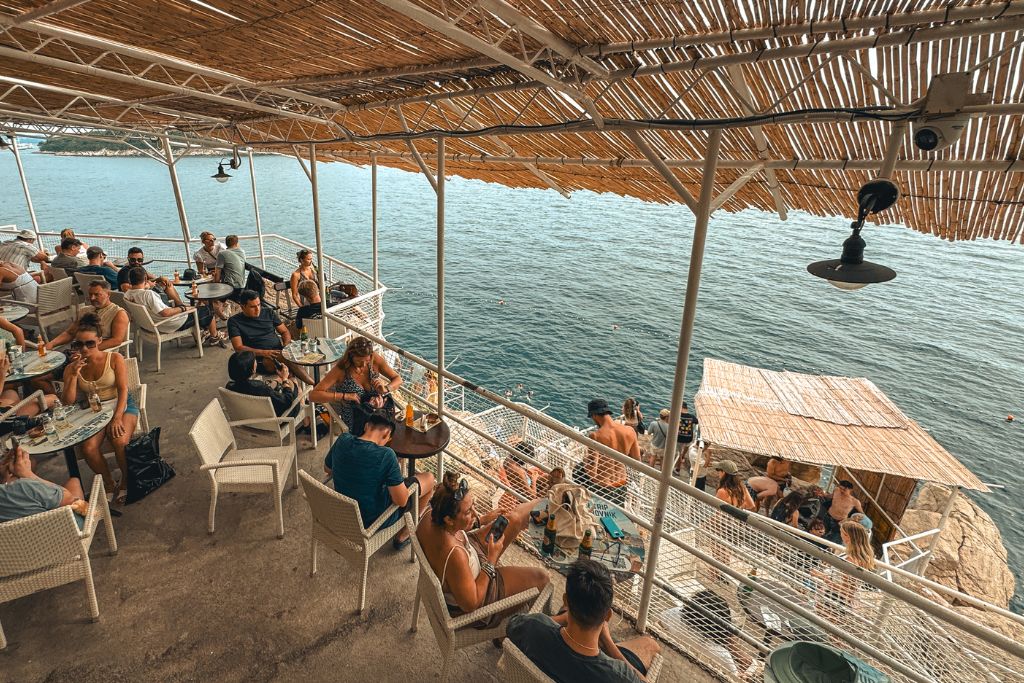 People sitting at small tables on a terrace along the cliffs, overlooking the Adriatic sea, with a thatched roof.