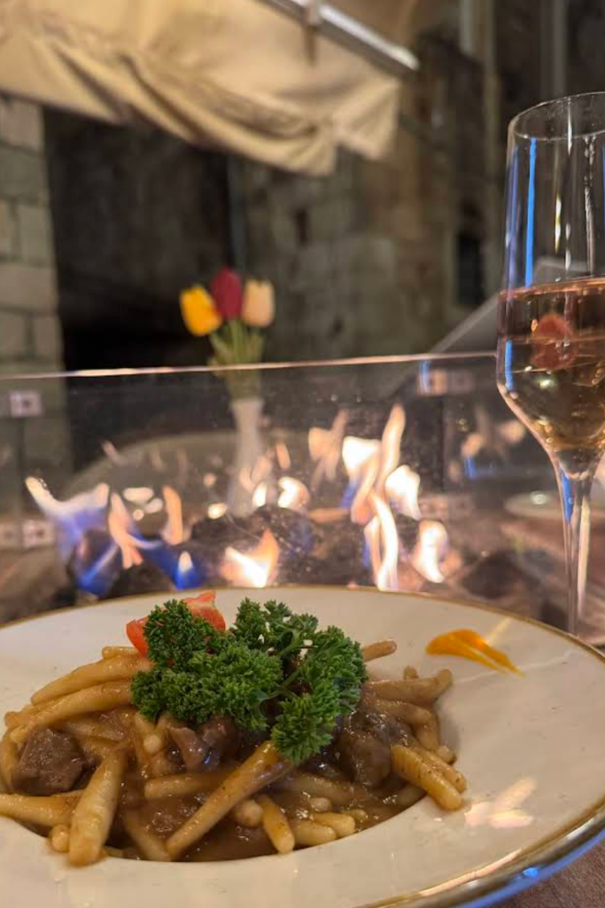 A plate of pasta on a table with a glass of wine and flames in the background.