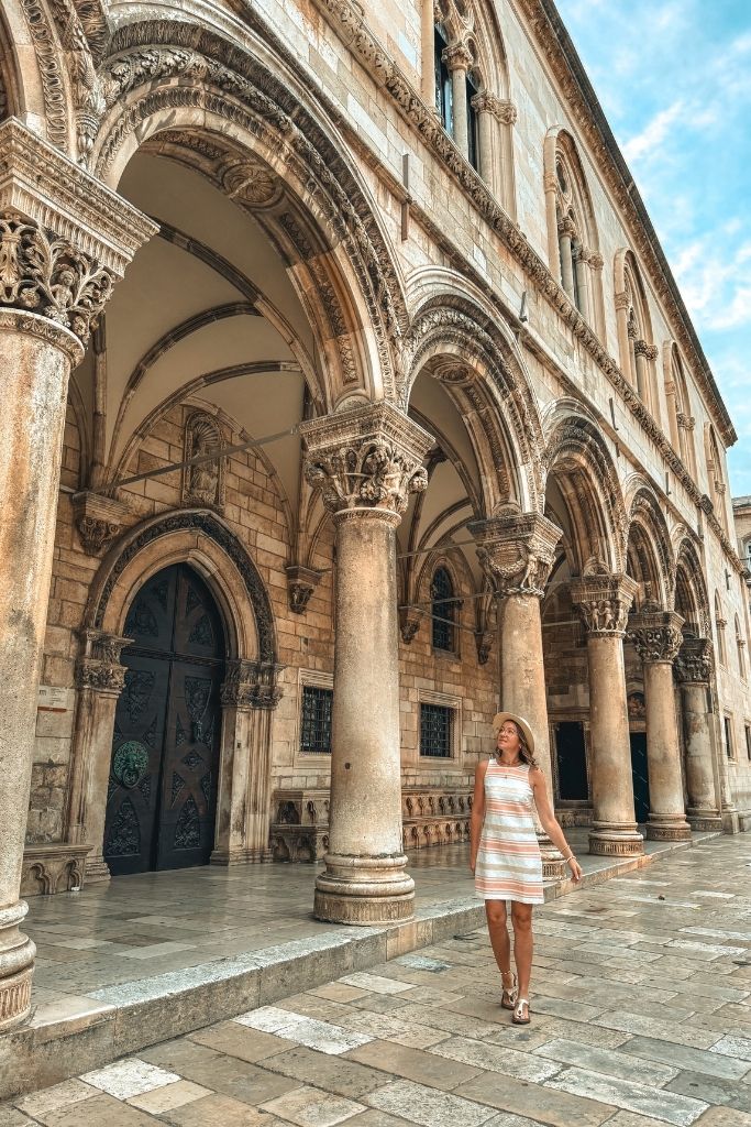 Kate walks by a gothic building with columns in Old Town Dubrovnik as she looks up at the architecture.