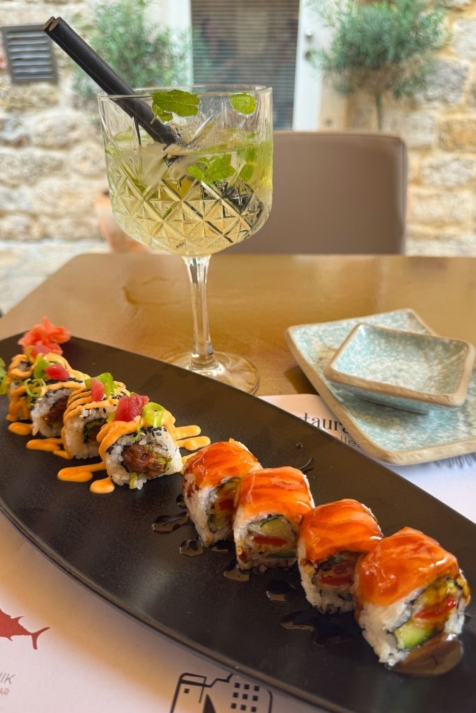 Table setting with a sushi roll on a long black plate and a Hugo Spritz in the background.