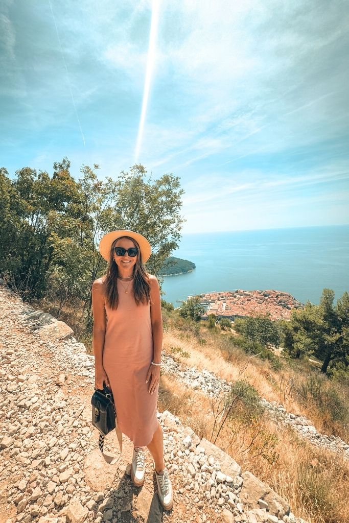 Kate stands in a pink dress and sneakers on a gravel trail that winds down towards the old town of Dubrovnik.