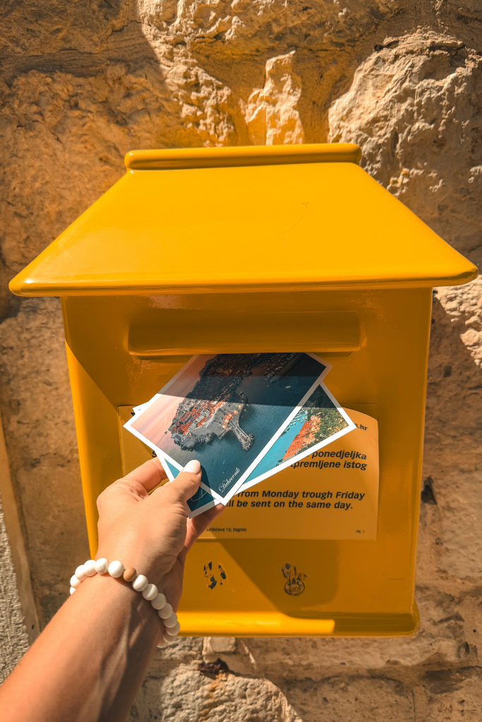 Kates hand dropping two postcards into a yellow mail box in Dubrovnik. 