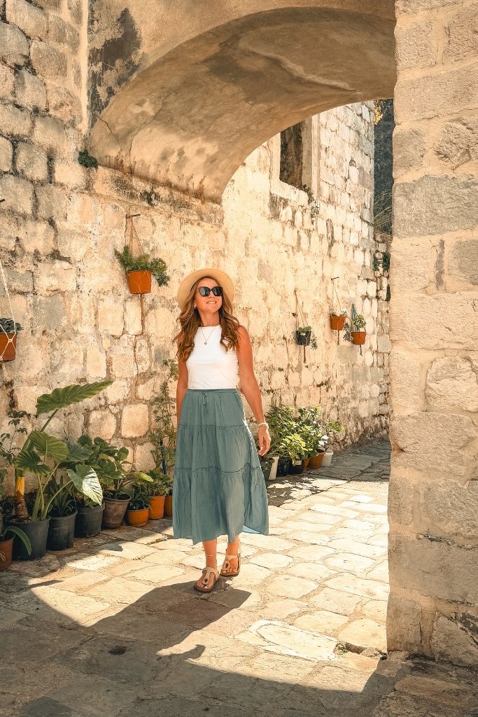 Kate walks through a narrow stone walled street with an arch overhead and colorful planters hanging on the wall.