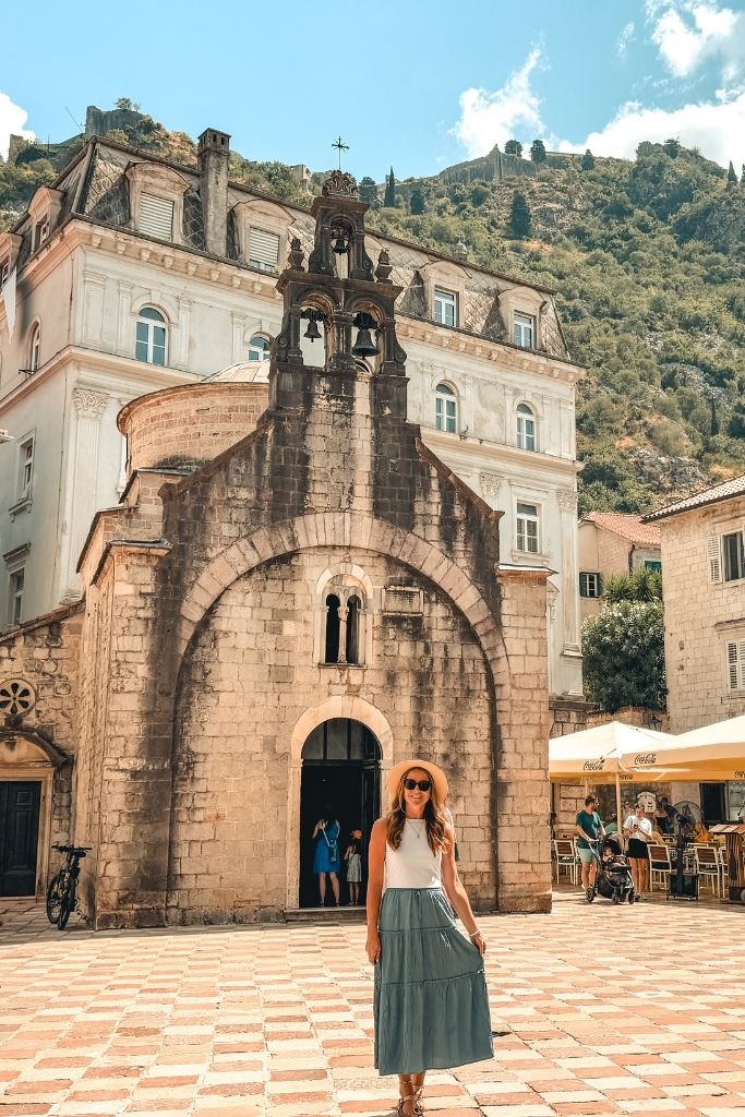 Kate walks through a square in Kotor with a small church and mountains in the backdrop.