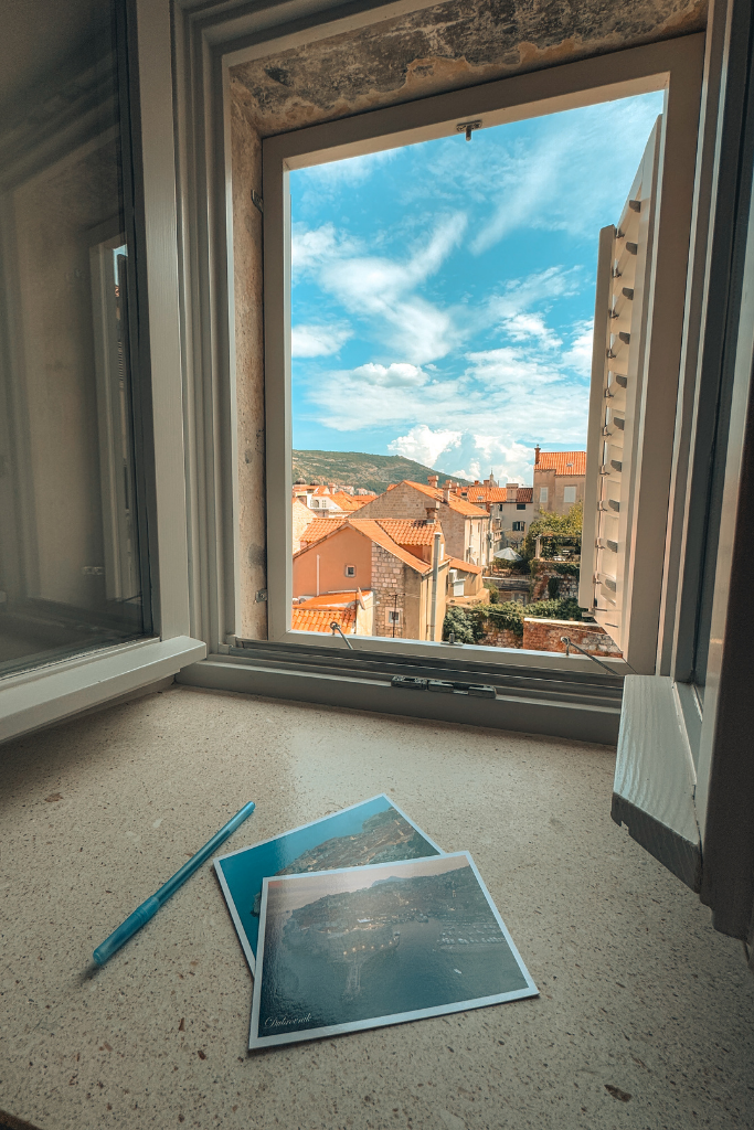 Two postcards sitting on a window seal with a view of Dubrovnik's old town. 