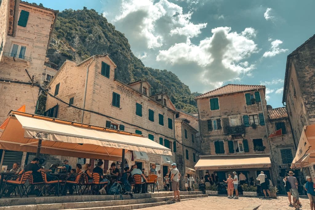 People enjoy lunch and drinks at a sunny cafe tucked inside the charming stone alleyways of Kotor Old Town beneath Montenegro’s dramatic mountain cliffs.