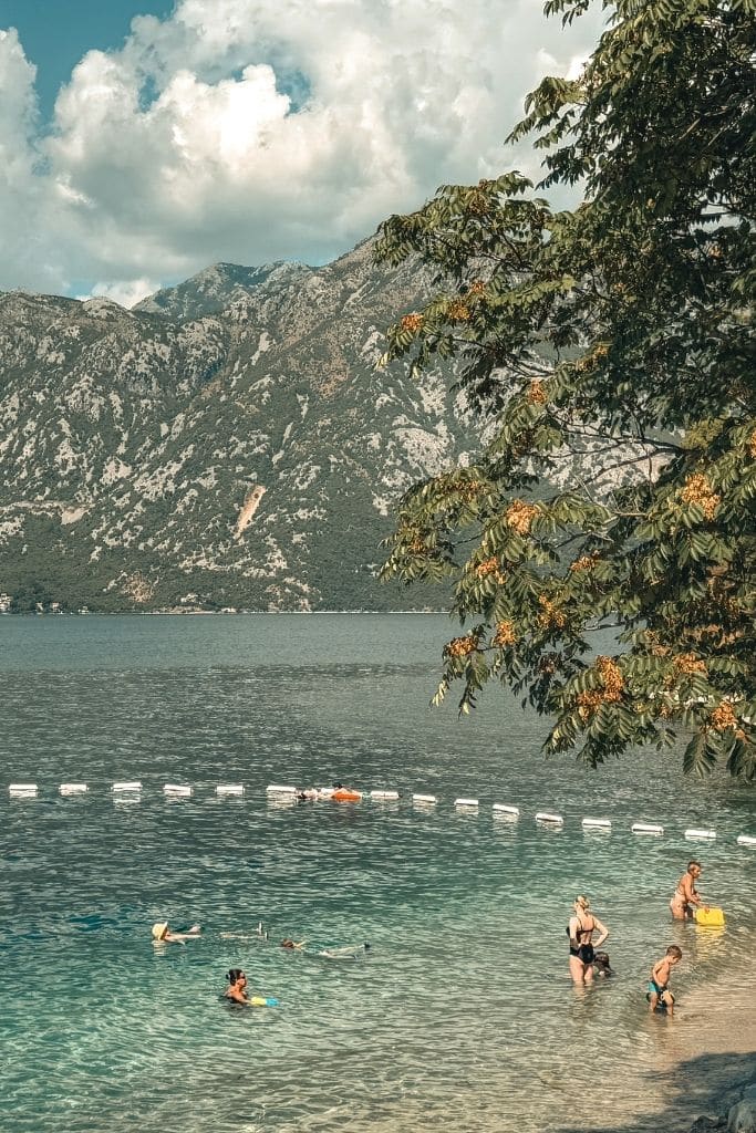 People swimming in the crystal-clear waters of the Bay of Kotor beneath Montenegro’s dramatic mountain scenery on a beautiful summer day.