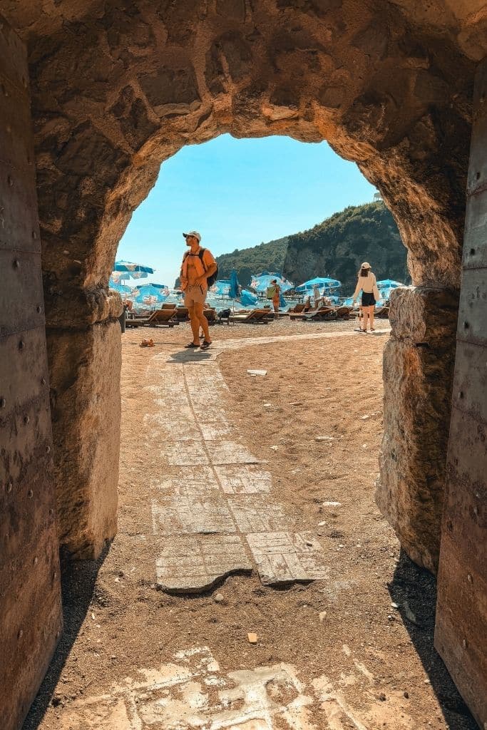 A stone archway in Budva’s Old Town opens up to a sunny beach scene along Montenegro’s beautiful Adriatic coastline.