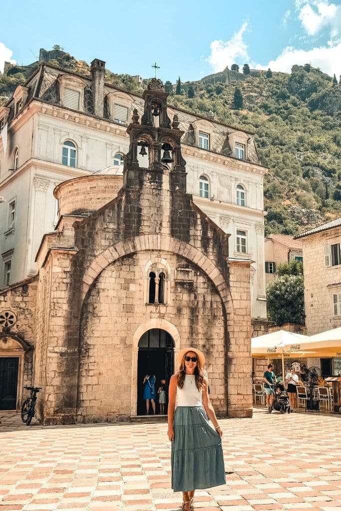 Kate explores one of Kotor Old Town’s charming historic squares, standing in front of a beautiful stone church beneath Montenegro’s dramatic mountain scenery.