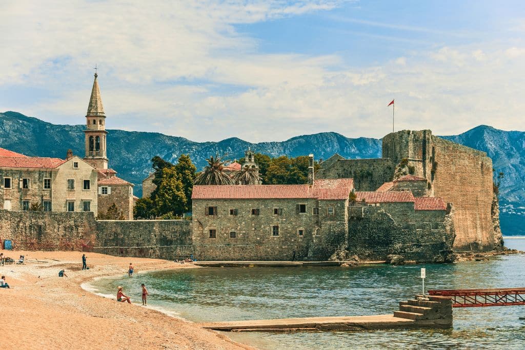 The fortified seaside walls of Budva’s Old Town sit along Montenegro’s Adriatic coastline, surrounded by mountains, historic stone buildings, and a quiet beach.