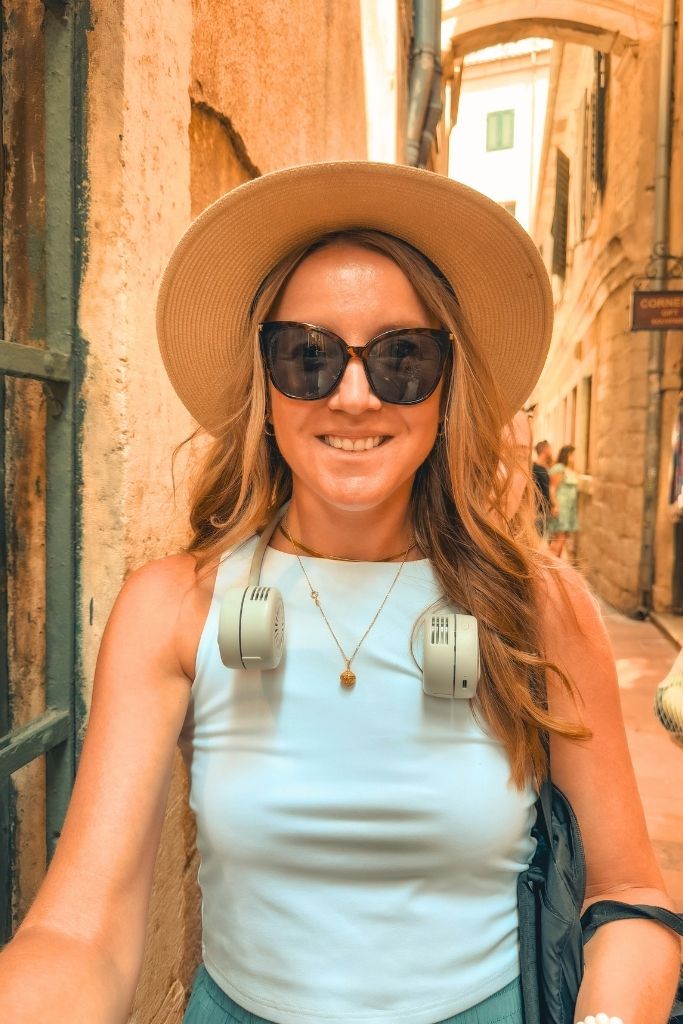 Kate smiles while standing in a narrow stone alleyway in a European old town, wearing a wide brim hat, sunglasses, a sleeveless white top, and a portable neck fan around her neck on a hot summer day.