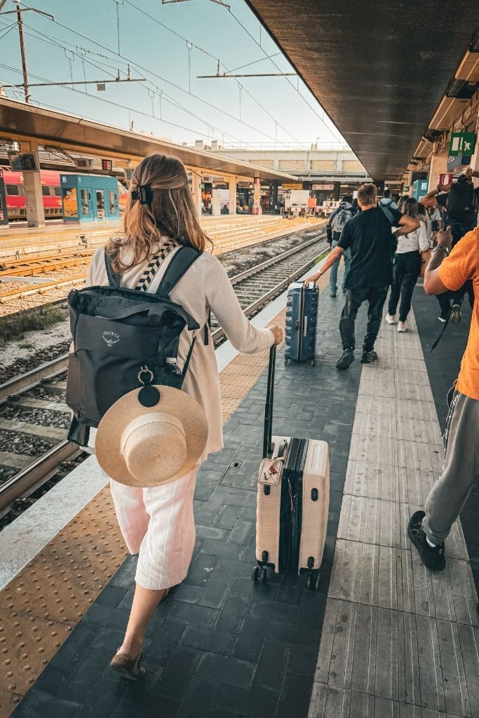 Kate walks through a busy European train station carrying a suitcase and backpack, dressed in lightweight travel clothes with a wide brim hat clipped to her bag for summer travel in Europe.