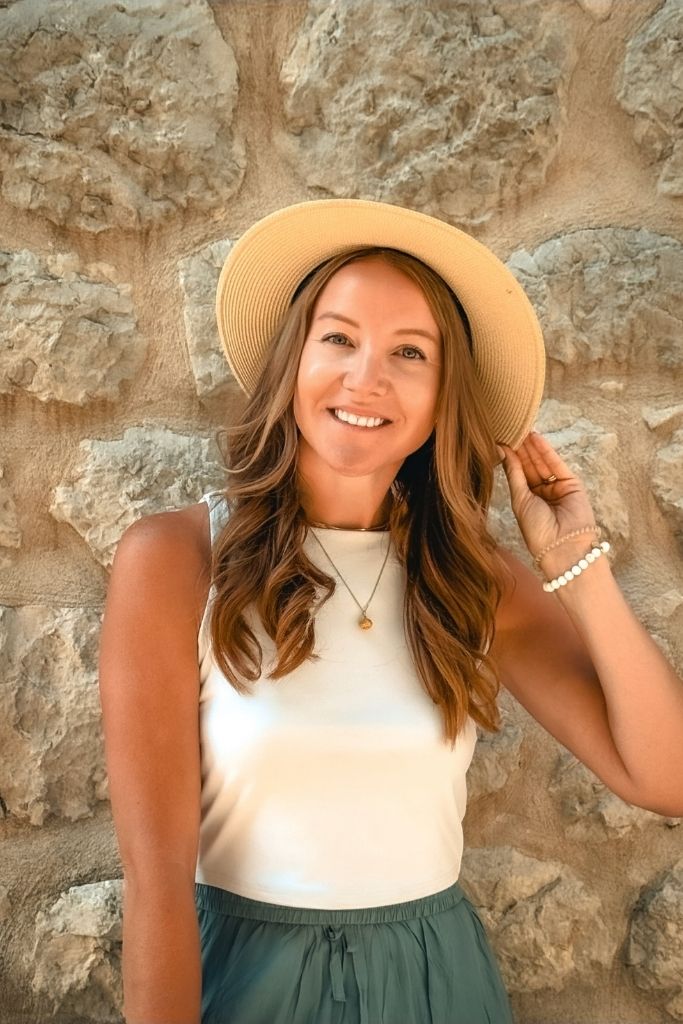 Kate smiles in front of a stone wall wearing a lightweight sleeveless top, flowy maxi skirt, and wide brim hat.