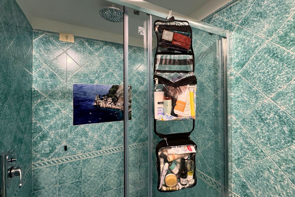 A hanging travel toiletry bag filled with skincare, sunscreen, and beauty essentials is organized inside a small European hotel bathroom.