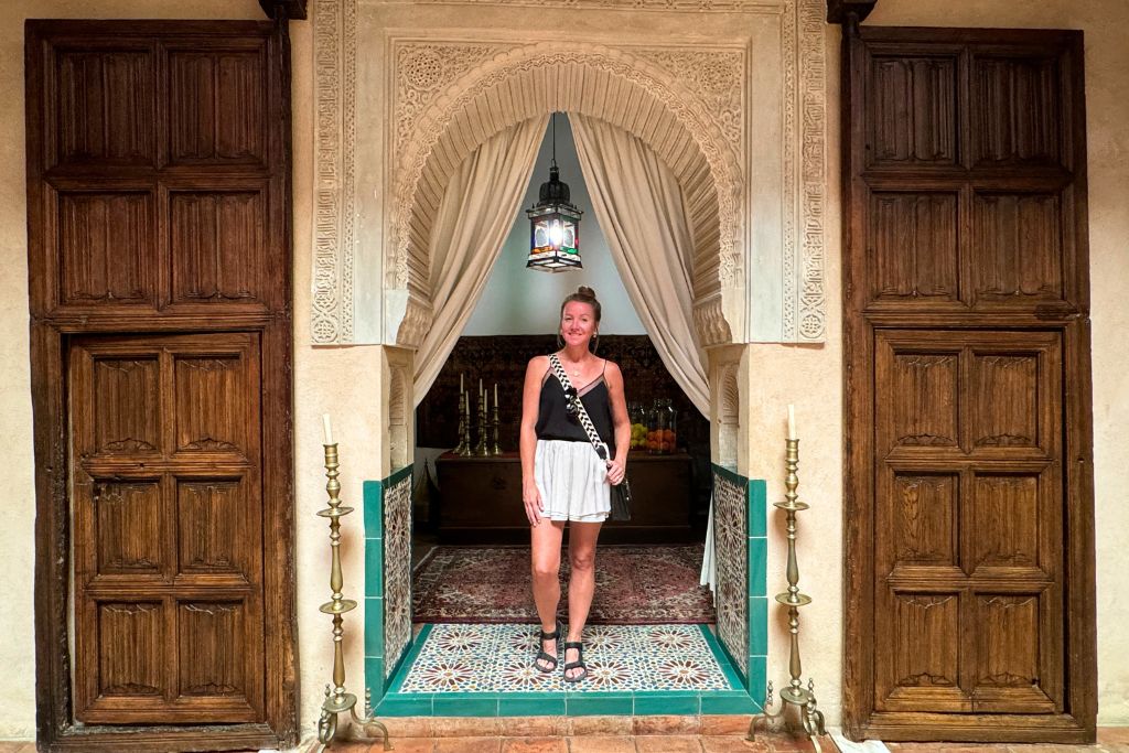Kate wearing a simple black tank top, flowy shorts, comfortable walking sandals, and a crossbody bag while exploring a beautiful historic hotel in Europe during summer.