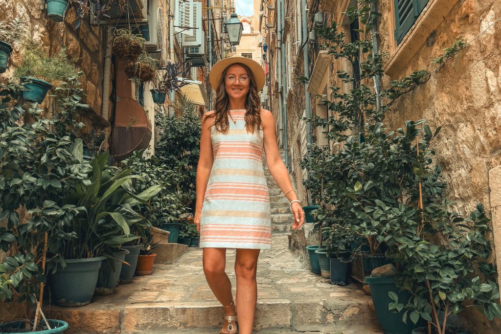 Kate wearing a lightweight striped summer dress, comfortable sandals, and a sun hat while walking through a charming narrow alley lined with stone walls and lush greenery in Europe during summer.
