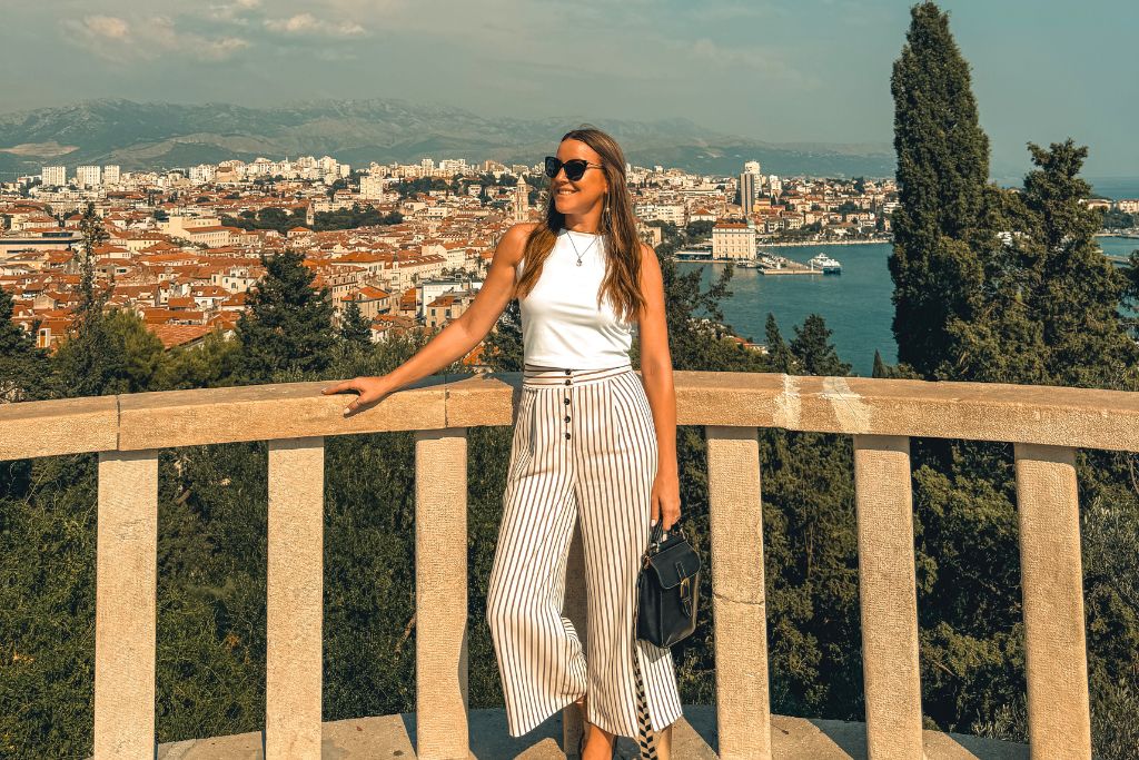 Kate wearing a white sleeveless tank top, striped wide-leg pants, sunglasses, and a crossbody bag while overlooking a scenic European coastal city in summer from a stone terrace viewpoint.