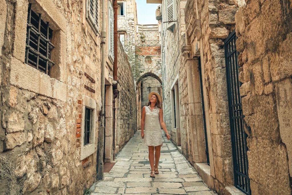 Kate wearing a lightweight neutral-colored linen shirt dress, comfortable walking sandals, and a sun hat while exploring a narrow stone alley in a historic European town during summer.