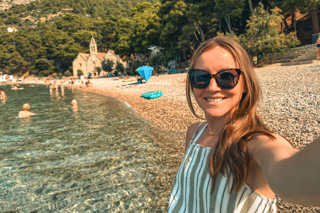 Kate wearing a striped lightweight summer top, sunglasses, and simple jewelry while taking a selfie along a crystal-clear beach in Europe during summer.