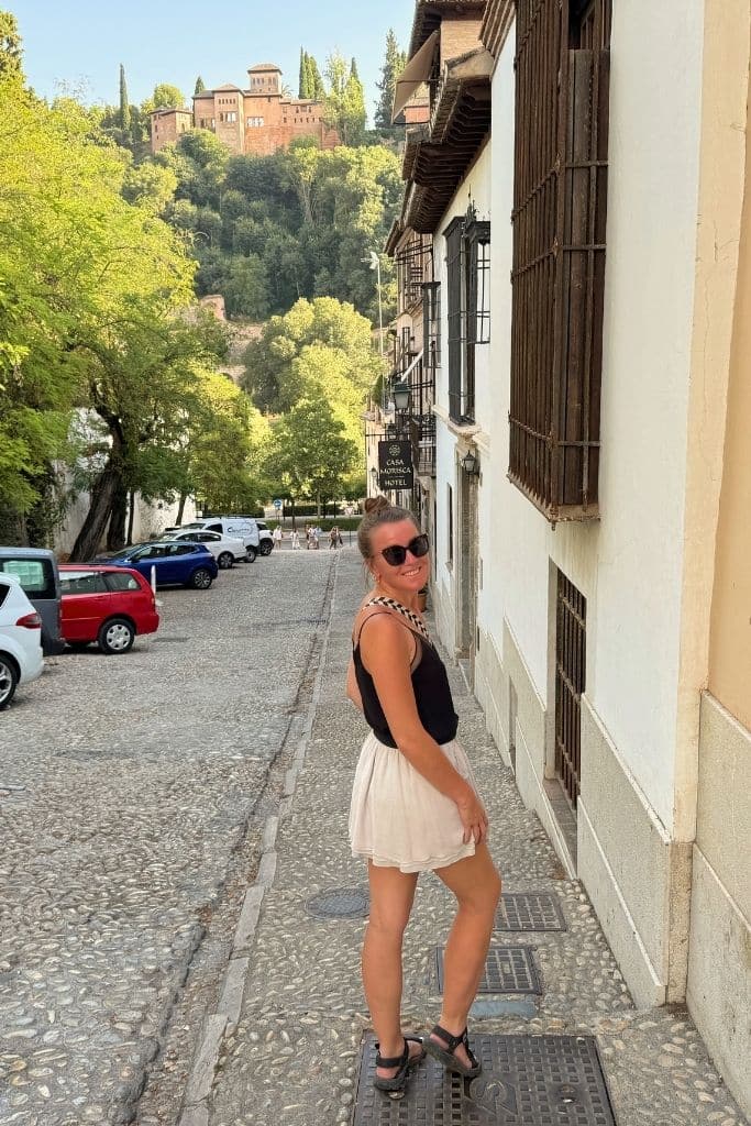 Kate wearing a simple black tank tow, short flowy skirt, and comforabel walking sandals on a cobblestone street in Europe in Summer.
