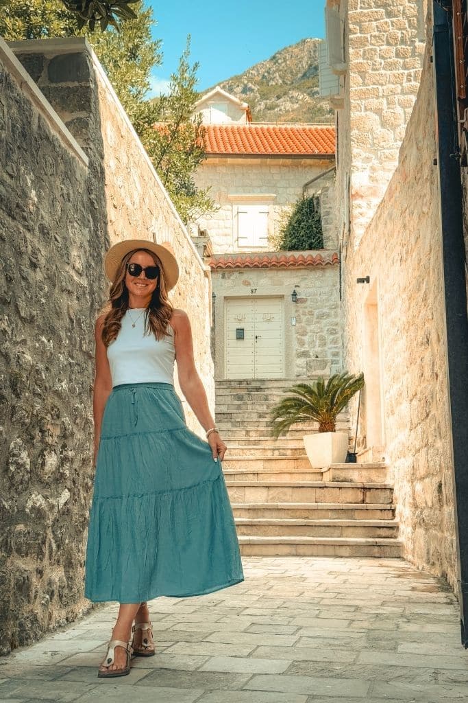 Kate in a blue flowy maxi skirt, wide brim hat, and comfortable sandals, waking down a narrow streen in Europe in Summer.